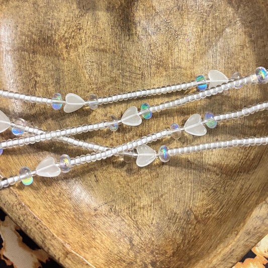 Glow in the dark waist beads with frosted heart charms and translucent white seed beads displayed on a wooden heart tray, glowing bright green in darkness
