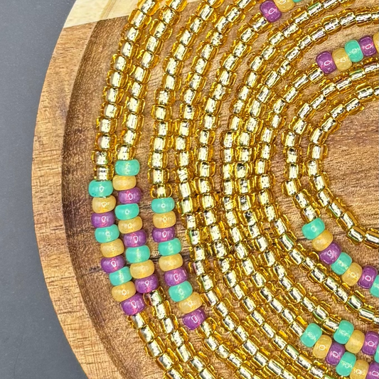 Gold waist beads with accents of teal, plum, and honey seed beads arranged in colorful clusters on a wooden display.