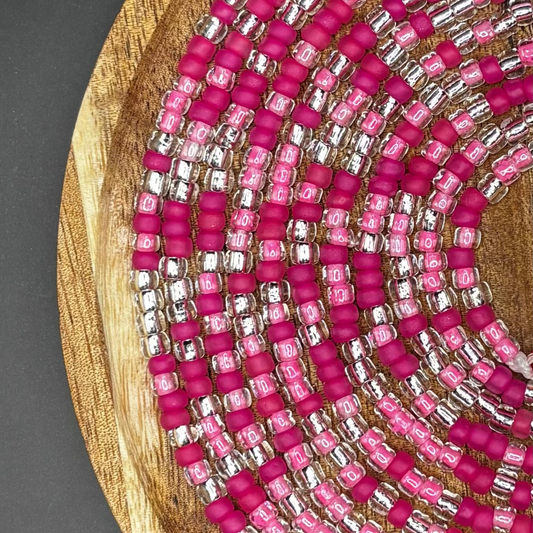 Pink and silver waist beads arranged in a spiral on a wooden tray, featuring bright fuchsia matte seed beads, translucent pink-lined beads, and metallic silver accents.