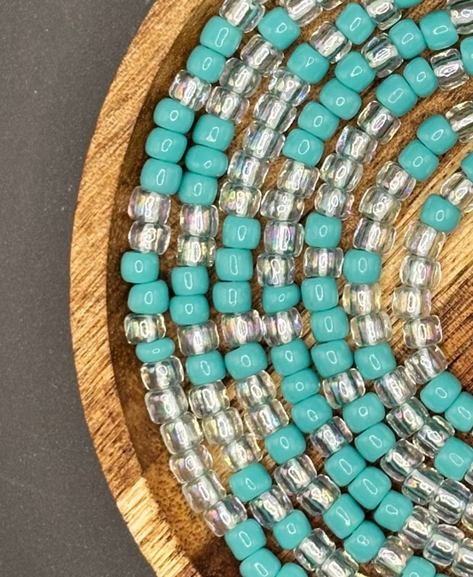 Turquoise and clear iridescent waist beads arranged on a wooden tray, featuring soft aqua seed beads paired with luminous crystal accents from Waisted by Design.